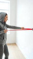 Young athletic woman in gray hoodie doing resistance band exercise at home near window for strength training and fitness workout.