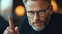 Mature bearded businessman in glasses making pointing gesture during conversation, close-up portrait with bokeh background lighting effects.
