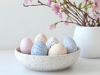 Pastel speckled Easter eggs in ceramic bowl with cherry blossom branches in white vase, minimal spring holiday composition on white background.