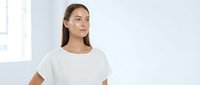 Young woman with long brown hair and light skin tone wearing a white casual t-shirt, looking thoughtfully to the side in a bright, minimalist interior setting for lifestyle and fashion content.