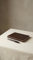 Closed brown leather notebook and black pen on beige linen table with neutral taupe wall background, minimalist workspace setup for business, office, and stationery design projects.