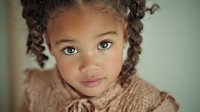 Young mixed-race girl with curly hair wearing beige knitted sweater looking at camera with bright eyes in soft natural lighting for family portraits.