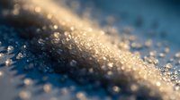 Macro close-up of sparkling frost crystals and ice particles glistening on a frozen surface with warm golden sunlight and soft blue background.