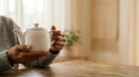 Young adult with medium brown skin tone pouring tea from ceramic teapot into cup at wooden table by window, wearing cozy sweater, ideal for beverage, lifestyle, and wellness content.