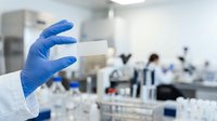 Laboratory technician wearing blue latex gloves holding test tube in modern research facility with blurred colleagues and equipment in background for scientific study.