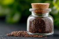 Black mustard seeds in small glass jar with cork lid on dark background against green bokeh. Aromatic spice and seasoning ingredient for cooking.