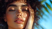 Young mixed race woman with curly hair enjoying sunlight, eyes closed with peaceful expression, natural makeup and glowing skin against blue sky background.