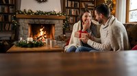 Young couple sharing intimate moment by fireplace during Christmas holidays, man giving gift to surprised woman in cozy living room with festive decorations.