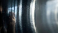 Young adult male with medium skin tone, wearing dark clothing, standing behind translucent plastic or glass with water droplets, creating a moody, atmospheric effect for conceptual and editorial use.