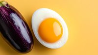 Fresh raw eggplant and sunny side up fried egg on vibrant yellow background, minimalist food composition with contrasting colors and textures.