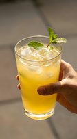 Refreshing yellow citrus cocktail with ice and fresh mint garnish held in hand against neutral background, suitable for beverage, hospitality, and summer drink marketing.