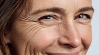 Close-up of mature woman with natural smile showing laugh lines and facial wrinkles against white background, representing aging gracefully.