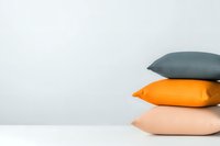 Minimalist composition of stacked decorative pillows in gray, orange, and peach colors against white background, showcasing modern interior design elements.