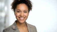 Young African American businesswoman with curly hair smiling confidently in professional blazer against blurred office background for corporate marketing.
