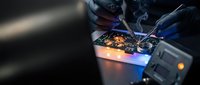 Electronics technician in blue gloves soldering circuit board components with precision tools in professional repair workshop environment.
