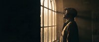 Young African American woman in profile standing by large window with dramatic lighting creating shadows and warm golden tones in interior space.