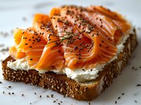 Fresh smoked salmon slices on multigrain bread with cream cheese spread and black sesame seeds, close-up view of gourmet open sandwich on white surface.