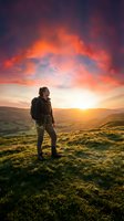 Hiker with backpack standing on grassy hillside at sunset, gazing at dramatic orange and purple sky over rolling mountains and valleys below.