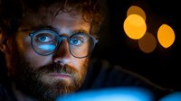 Young bearded man with glasses illuminated by blue light from screen working late at night, orange bokeh lights in background creating moody atmosphere.