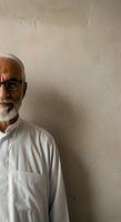 Senior man with white hair and beard wearing eyeglasses and white shirt standing against textured beige wall. Portrait suitable for healthcare, business, professional services, and lifestyle editorial use.