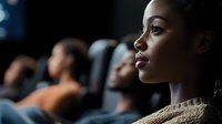 Young African American woman watching intently in dark theater audience, profile view showing elegant features and pearl necklace against blurred background.