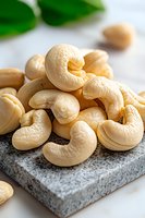 Fresh raw cashew nuts on gray stone board with green leaves in background, macro food photography showing texture and natural cream color.