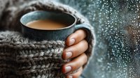 Cozy winter morning with steaming coffee mug held by female hands wearing knitted sweater against frosty window with water droplets creating warm atmosphere.