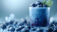 Fresh blueberry smoothie in glass garnished with whole berries and mint leaves on blue background with scattered berries, refreshing summer drink.
