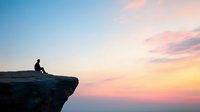 Silhouette of person sitting alone on cliff edge against dramatic sunset sky with pink and orange clouds, contemplative solitary moment in nature.