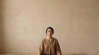 Young woman with dark hair and South Asian ethnicity wearing a beige linen shirt against a neutral tan wall. Suitable for portrait, lifestyle, wellness, and corporate design projects.
