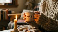 Cozy woman in knit sweater holding steaming mug with love inscription near burning candle, warm hygge home atmosphere.
