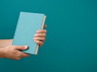 Two hands holding an open light blue book against a teal background, suitable for education, reading, and publishing concepts.