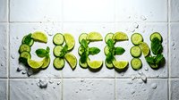 Word GREEN spelled with fresh cucumber slices, lime wedges, and mint leaves on wet white tile background with ice cubes.