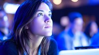 Young woman with thoughtful expression illuminated by blue light at conference or presentation, looking upward with anticipation.
