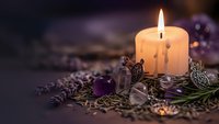 Burning candle surrounded by dried lavender flowers, amethyst crystals and decorative metal charms creating peaceful spa atmosphere for relaxation and meditation.