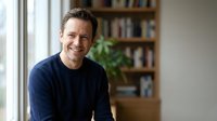 Middle-aged Caucasian man smiling warmly in modern home office with bookshelf background, wearing navy sweater for professional lifestyle content.
