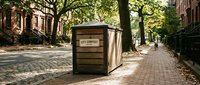 Vintage newspaper kiosk on tree-lined urban street with brick buildings and pedestrians in background, suitable for editorial and architectural stock use.
