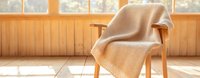 Cozy knitted beige blanket draped over wooden armchair by sunlit window with warm natural lighting streaming across hardwood floor. Minimalist home interior setting.