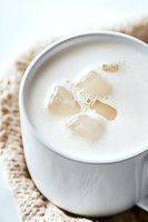 Creamy white hot drink with melting ice cubes in ceramic mug on knitted cloth, close-up view showing refreshing cold beverage with frothy surface.