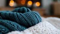 Cozy knitted teal sweater detail with soft white blanket against blurred warm bokeh lights background, creating atmospheric winter comfort mood.