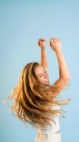 Happy young woman with flowing blonde hair dancing with arms raised against blue sky background expressing joy and freedom in summer lifestyle moment.