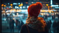 Young woman in red knit beanie looking at city lights at night, blurred bokeh background creates atmospheric mood, orange and teal color scheme, winter urban scene.
