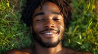 Young African American man lying in green grass with peaceful smile and closed eyes, enjoying nature and sunlight in relaxing outdoor moment.