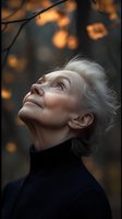 Senior woman with silver hair in black turtleneck gazing upward at autumn leaves, illuminated by warm golden light against dark background. Peaceful contemplative expression.