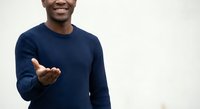 Young African American man in navy blue sweater making welcoming gesture with open hand, smiling warmly against neutral background for business presentations.