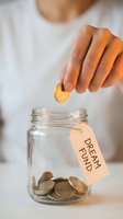 Close-up of person's hand dropping golden coin into glass jar labeled dream fund, symbolizing savings, financial goals, and wealth accumulation for future planning.
