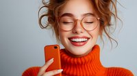Young woman with bright smile wearing orange turtleneck sweater and round glasses taking selfie with smartphone against gray background. Casual lifestyle portrait.
