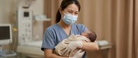 Asian female nurse in scrubs and medical mask holds newborn baby in hospital maternity ward, representing healthcare and neonatal care.