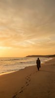 Solitary figure walking along golden beach at sunset with footprints in sand and dramatic orange sky reflecting on ocean waves for peaceful meditation concepts.