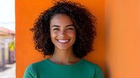 Young African American woman with natural curly hair and bright smile wearing green top against vibrant orange wall background, expressing joy and confidence.